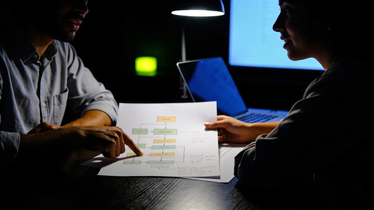 Duas pessoas conversando sobre um diagrama de processo impresso, à noite, iluminadas por uma luminária de mesa