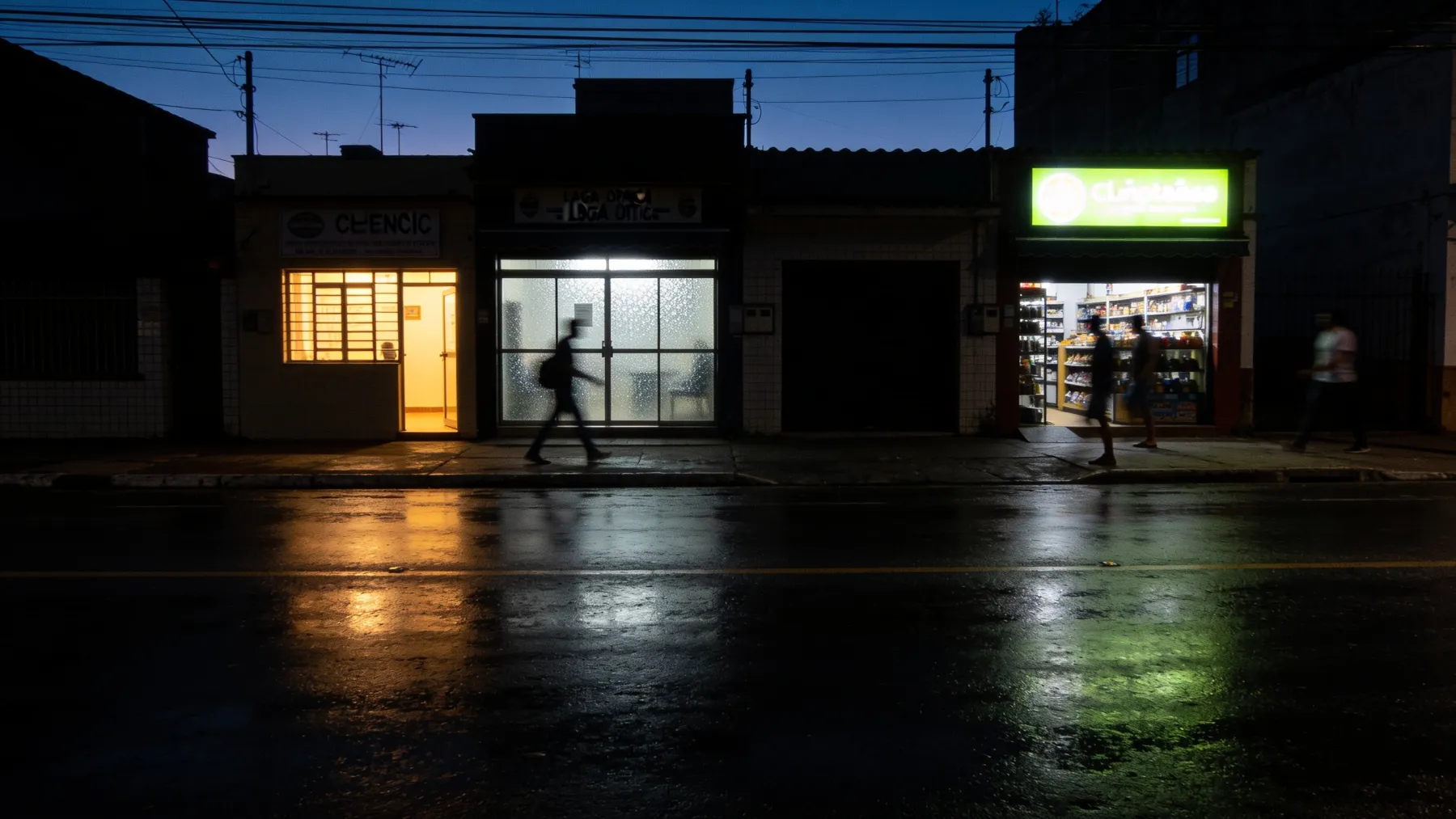Rua de comércio de bairro no fim da tarde, com vitrines acesas de uma clínica, um escritório e uma loja